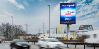 Im Vordergrund des Bilds sind verschwommen fahrende Autos wahrzunehmen. Der Fokus liegt auf der großen Werbetafel, die auf einem Mast über die Straße ragt. Es wird Werbung für den „ADVERTOWER" gezeigt. Im Hintergrund ist ein Gewerbegebiet zu sehen.