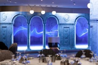 Eine Aufnahme einer Unilumin-LED-Wandinstallation auf dem Kreuzfahrtschiff „Spectrum of the Seas“. Abgebildet ist der Speisesaal, mit Blick auf die LED-Wände im Eingangsbereich. Im Vordergrund stehen chic eingedeckte Tische mit Sesseln.